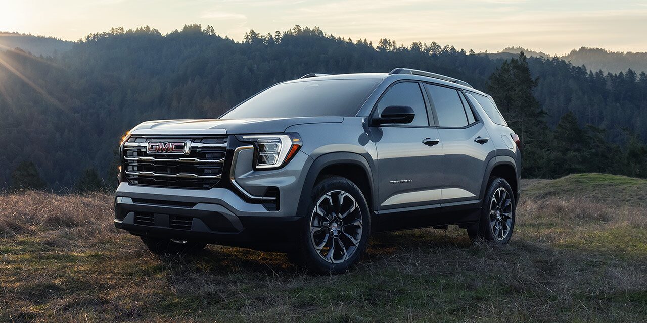 Front Three-Quarters View of the 2026 GMC Terrain Small SUV Parked in a Grass Field as the Sun Peaks Over the Tree Tops