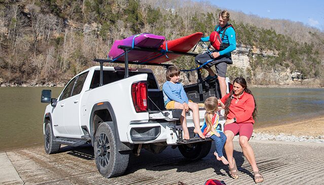 A Family of Four Sitting on the Tailgate of a White GMC Sierra Truck with Kayaks on the Roof Rack by a Lake