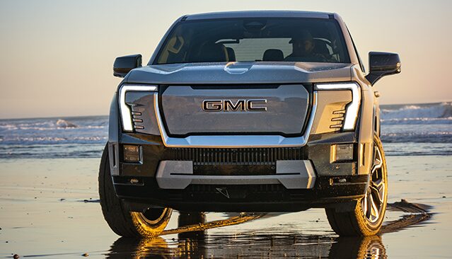 The Front View of a Gray GMC Sierra EV Truck Parked on a Sandy Beach at Sunset, with Ocean Waves in the Background