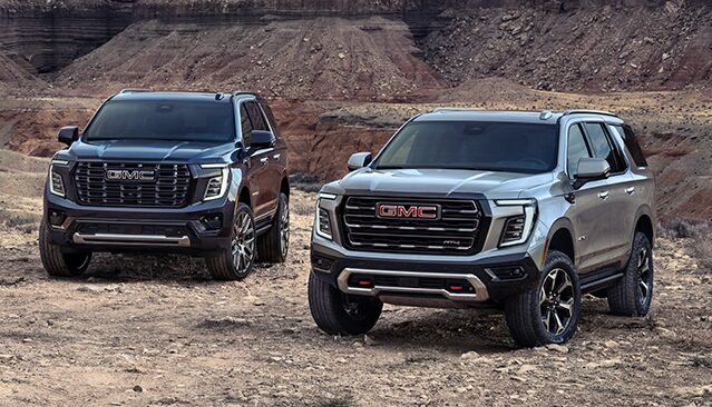 Two GMC Yukon SUVs, One Dark Gray and One Light Gray, Parked on a Rocky, Desert-like Terrain