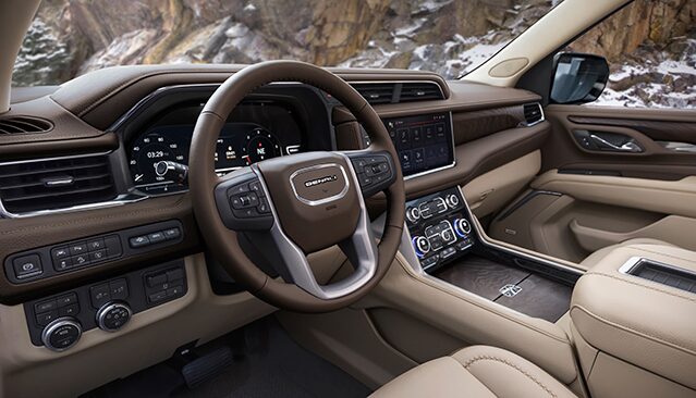 The Interior of a GMC Truck, Featuring a Brown Leather Steering Wheel, Dashboard, and a Large Central Touchscreen, with Snow-Covered Mountains Outside