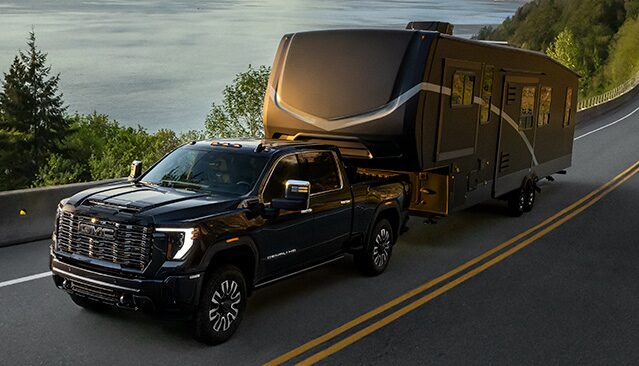 A Dark GMC Sierra HD Denali Pickup Truck Towing a Large Fifth-Wheel Camper on a Coastal Highway at Sunset