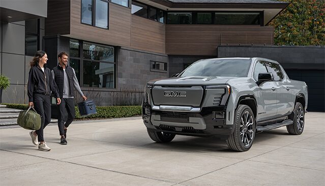 A Light Gray GMC Sierra EV Truck Parked In a Driveway in Front of a Modern House, with a Man and Woman Walking Toward It