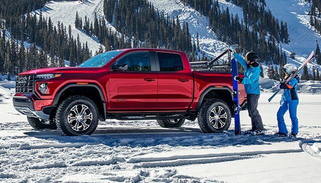 A Red GMC Canyon AT4X Pickup Truck Parked in a Snowy Landscape with Two People Loading Skis Onto a Roof Rack