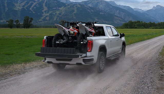 A White GMC Sierra Truck Driving on a Dirt Road with Two Dirt Bikes in its Bed and Mountains in the Background