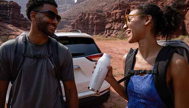 A Man and Woman Smiling and Holding a White GMC Water Bottle with a GMC SUV in the Background in a Desert