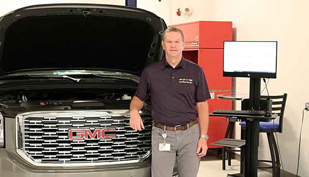 A Man Wearing a GMC Service Uniform Standing in a Garage With a GMC Truck and Diagnostic Equipment