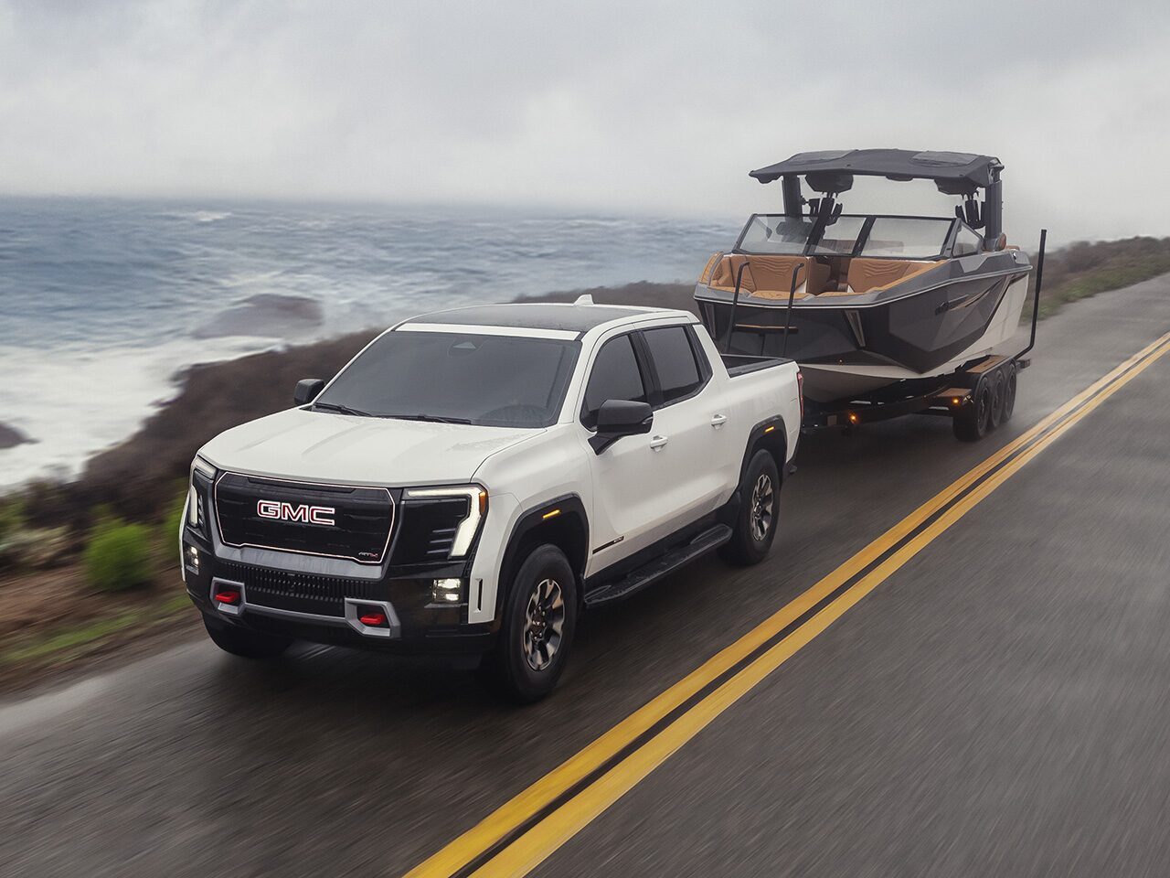 Three-quarters View of a White 2026 GMC Sierra EV Pickup Truck Towing a Boat near an Ocean Road