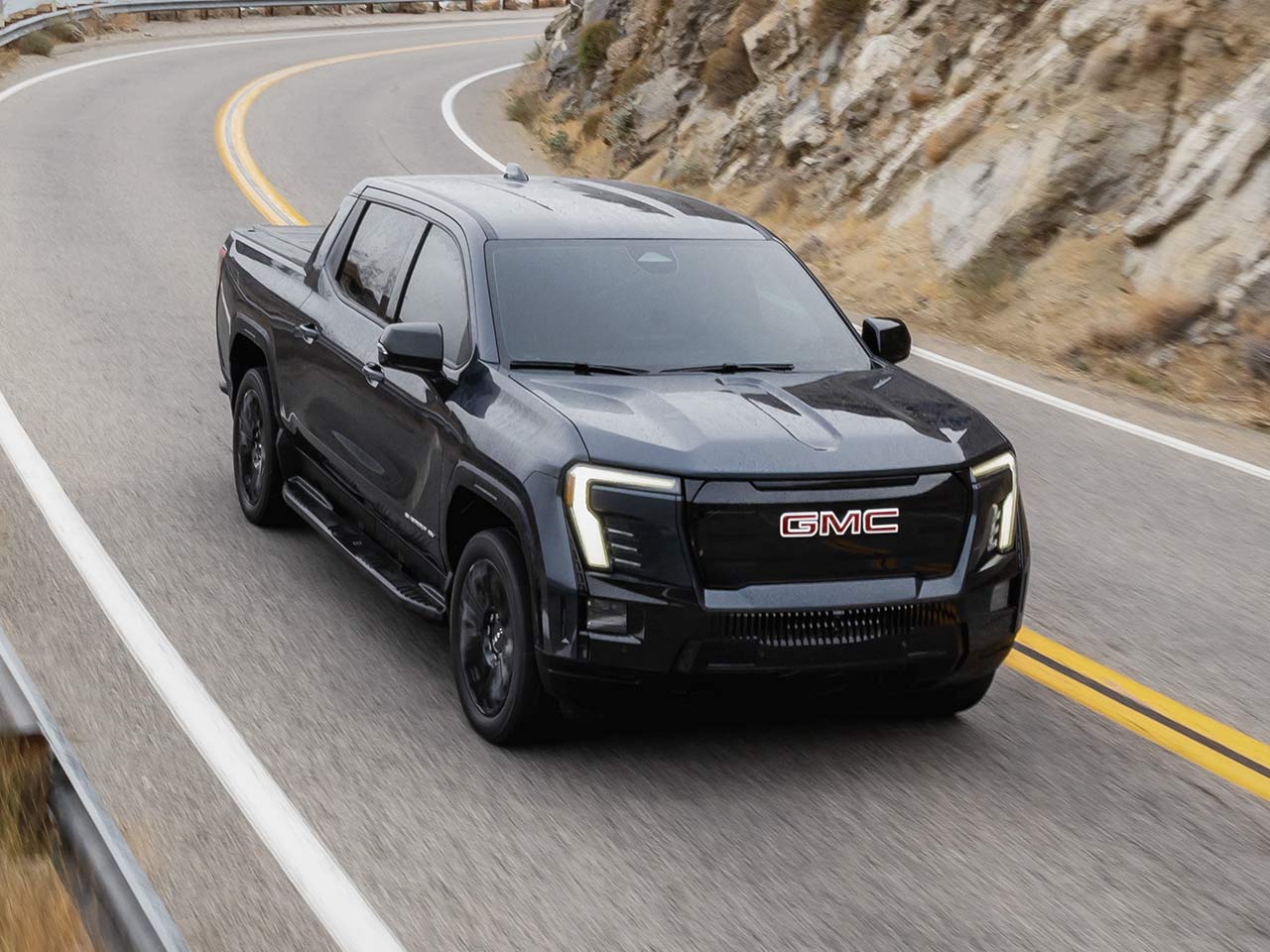 Three-quarters Passenger Side View of a Black 2026 GMC Sierra EV Driving Through a Mountain Road
