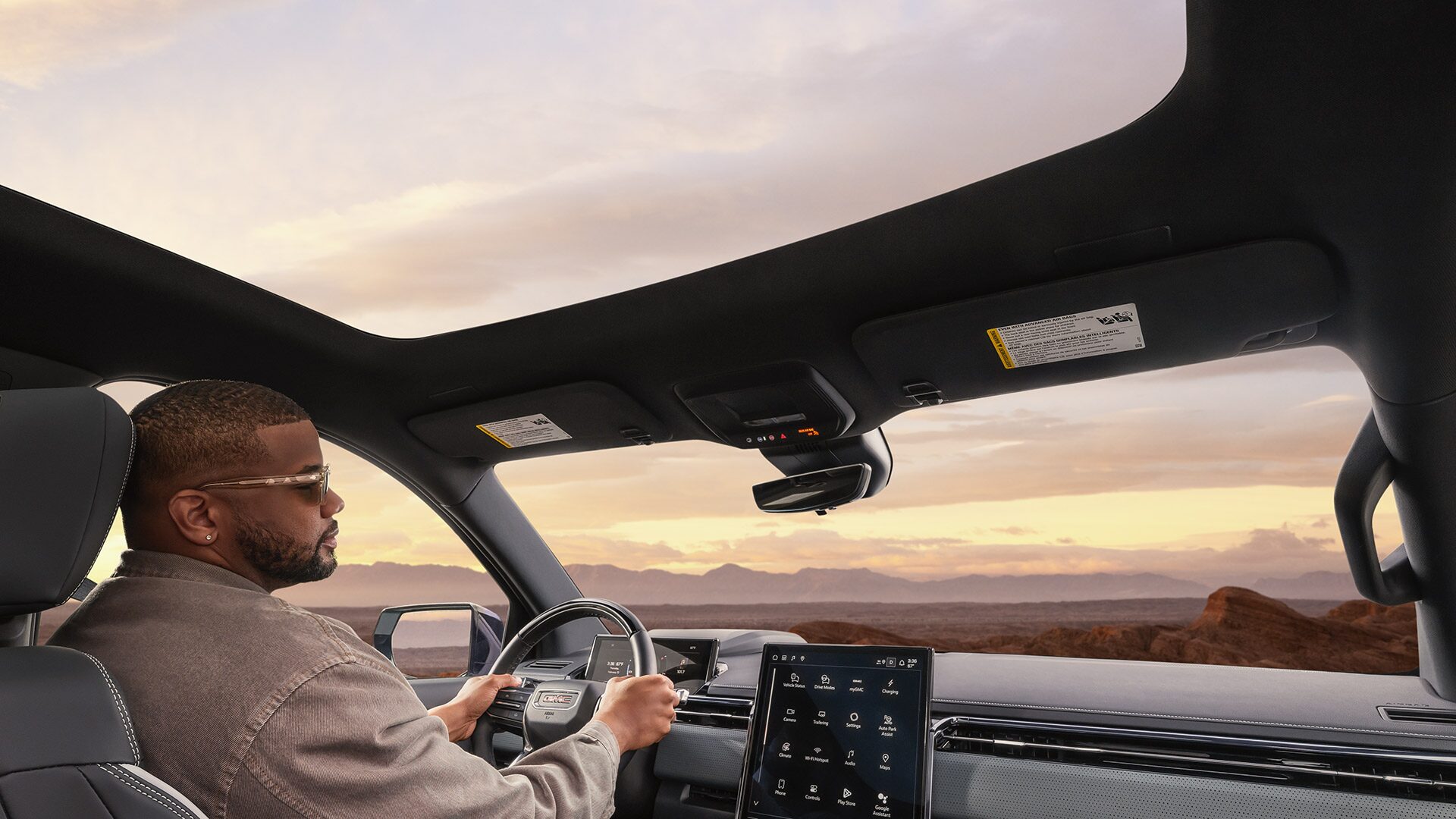 Back Seat View of a Man Driving the 2026 GMC Sierra EV with the Panoramic Sunroof Open During a Beautiful Sunset