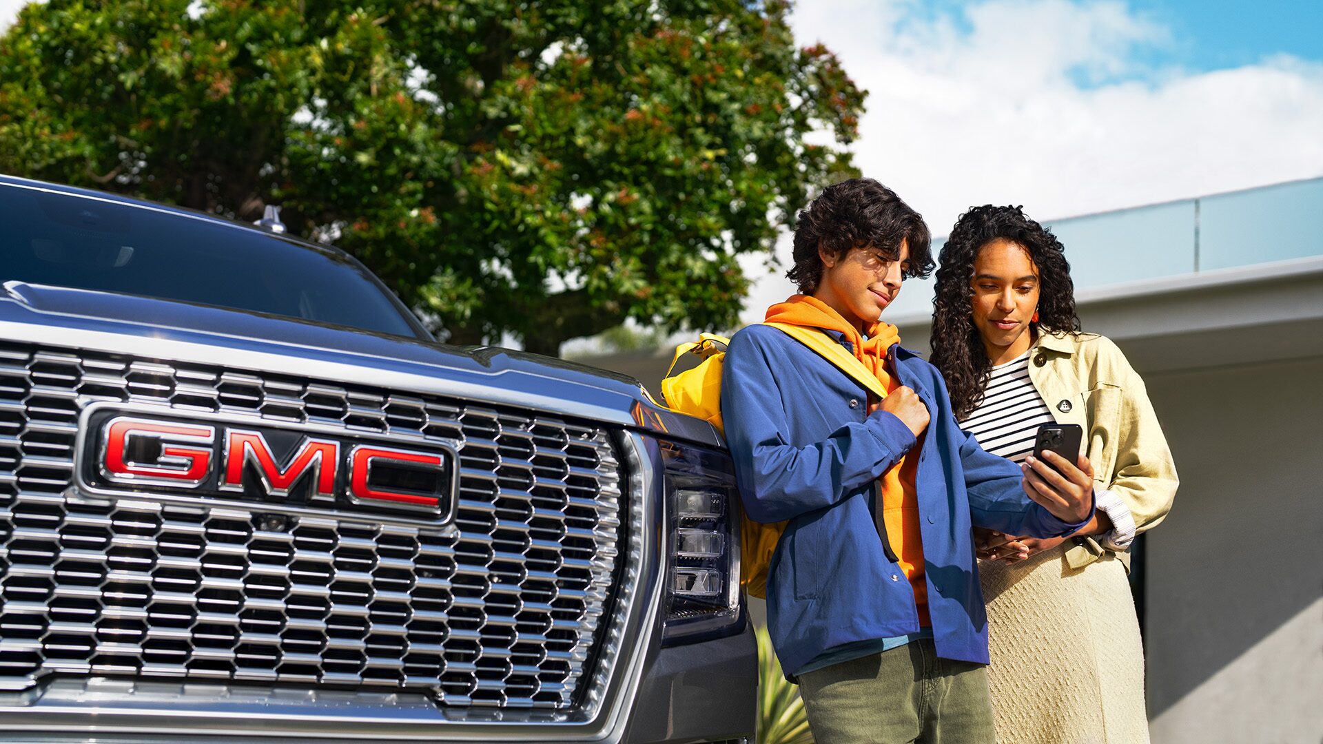 A smiling young man wearing a blue jacket and orange hoodie shows a smartphone to a woman standing next to him, next to the chrome grille and GMC logo of a dark SUV, checking information on how to upgrade an OnStar plan.